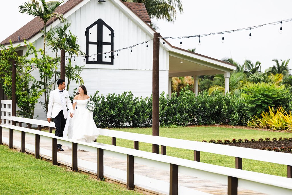 Leah and Jose walking beside the white fence and barn at The Arbor Estate
