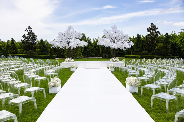 All-white outdoor ceremony design with clear chairs and flowering trees